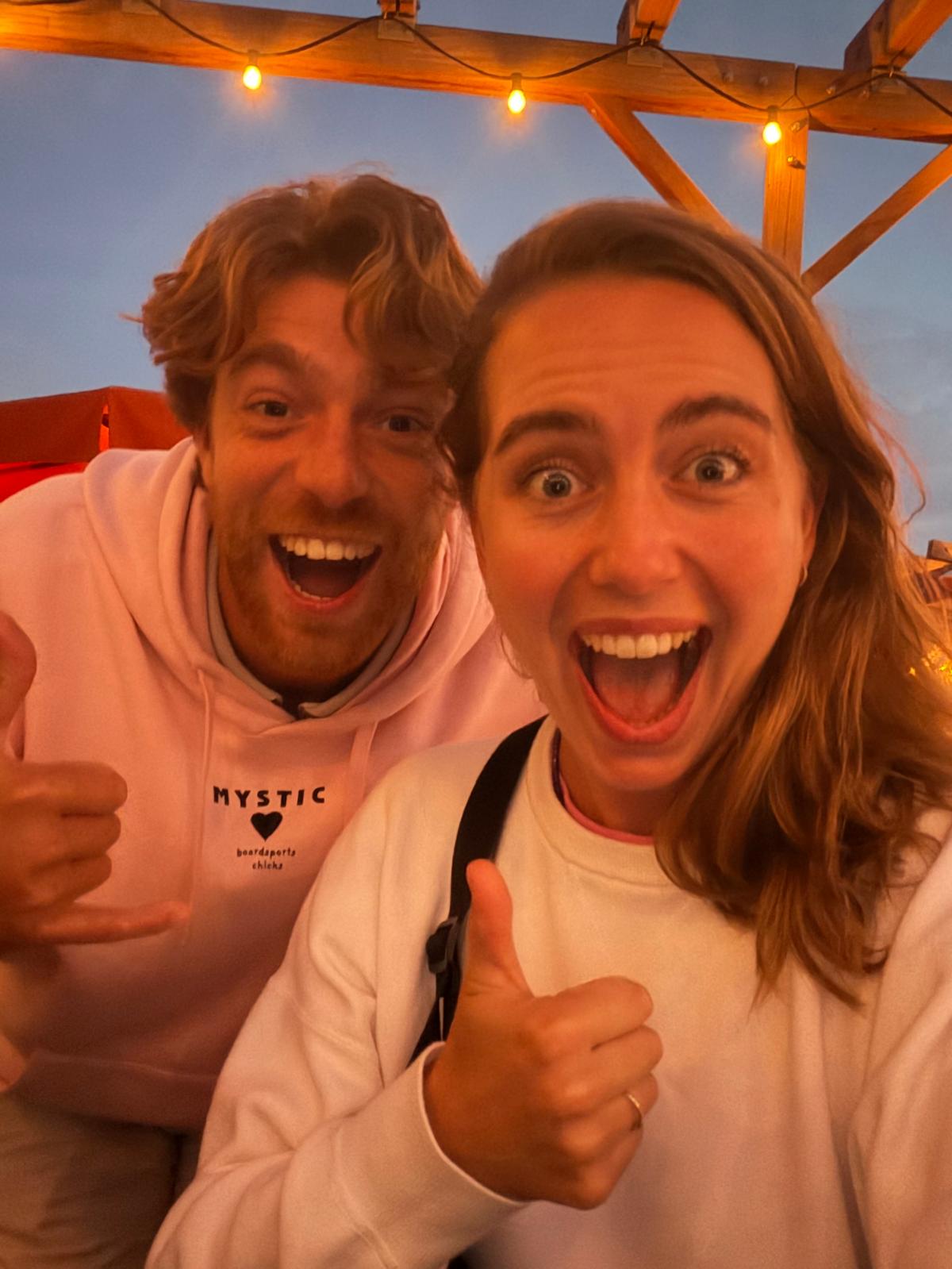 Jelle and Mieke on the beach at dusk, giving thumbs up under string lights