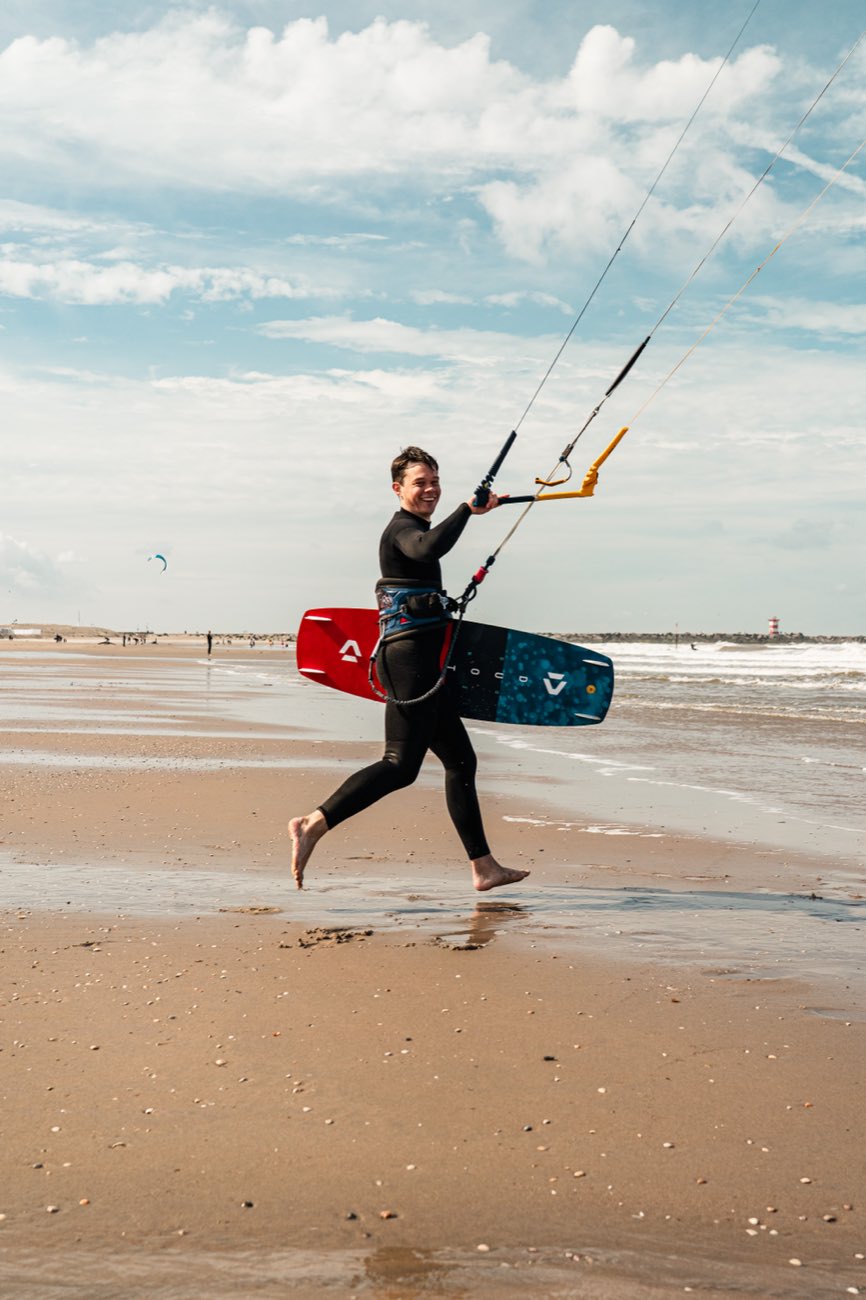 A kitesurfer running back along the beach with his board under his arm, grinning, kite still up